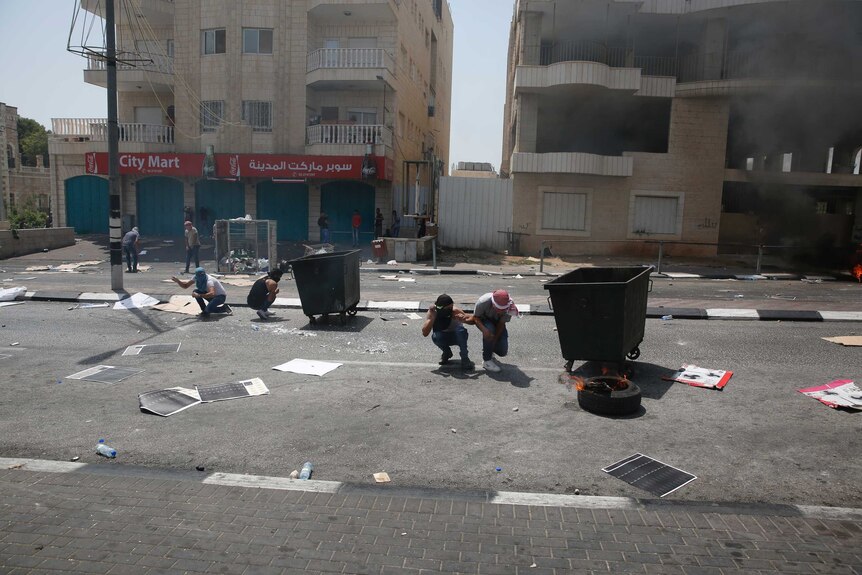 Young men with their faces covered kneel behind large bins on a road.