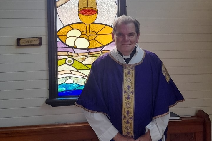 An Anglican priest dressed in white and blue robes stands in front of a stained-glass window