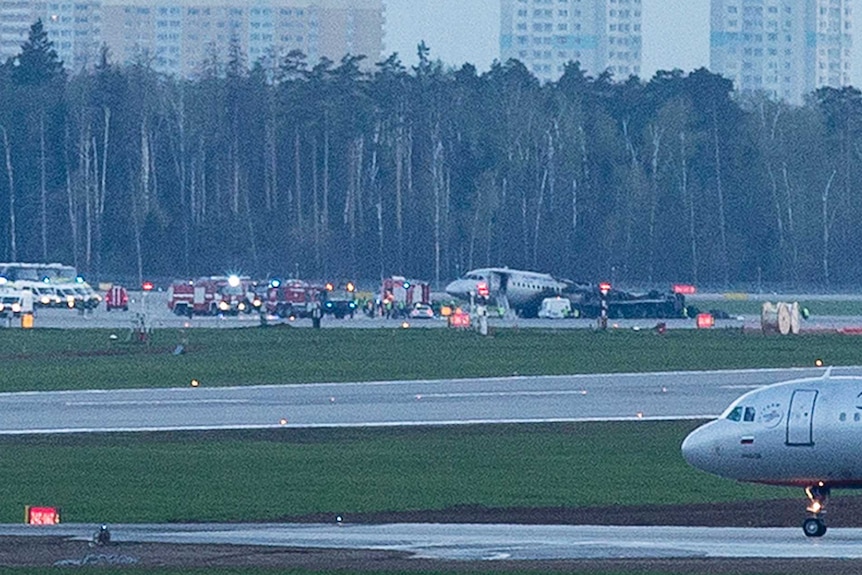 The charred back end of the plane seen at a distance as emergency vehicles surround the plane.