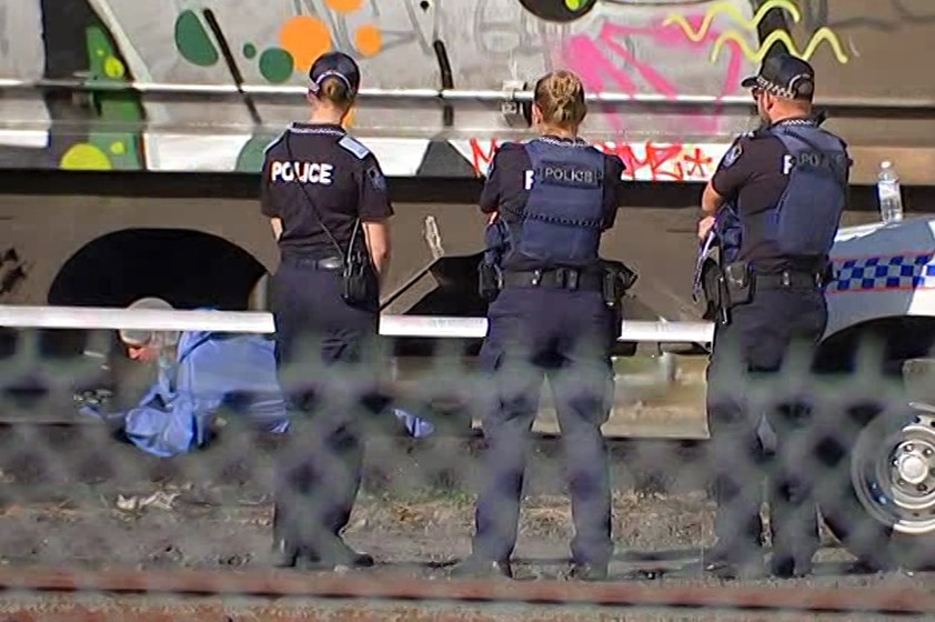 Forensic officer looks under a freight train with police officers alongside at Port of Brisbane