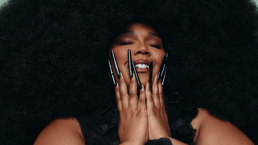 Lizzo has a huge afro haircut and enormous black fingernails. She holds her hands to her face and grins with her eyes closed.