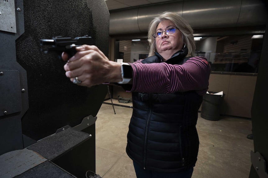 A woman in a puffy vest and glasses aims a gun at a shooting range