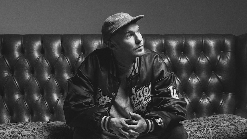 Black and white photo of Drapht sitting on a leather chesterfield couch wearing a baseball cap and Los Angeles Dodgers jacket