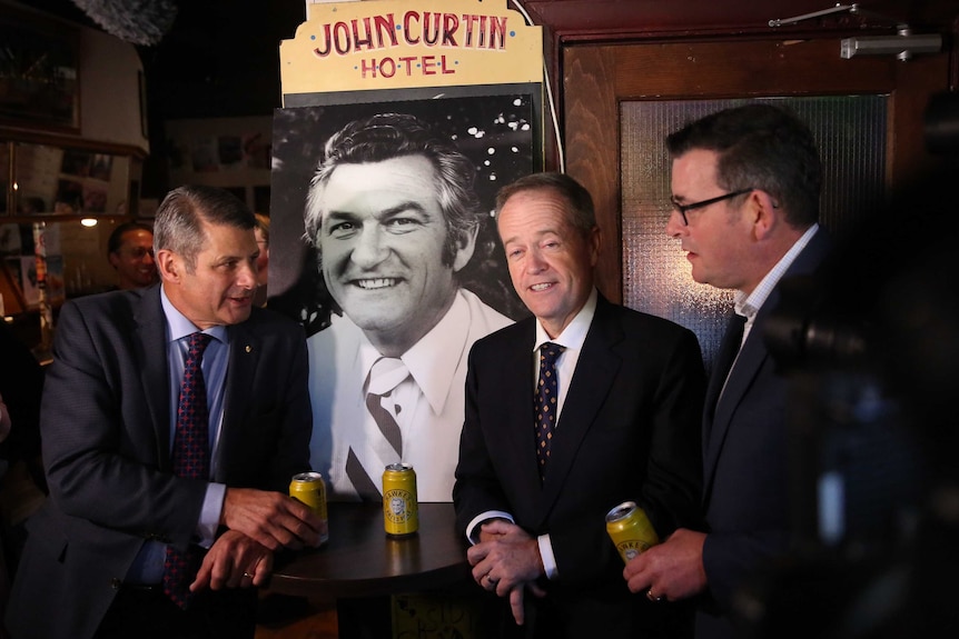 Steve Bracks, Bill Shorten and Daniel Andrews drink Bob Hawke beers as a stand around a photograph of the former PM