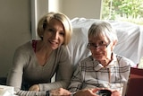 A middle-aged woman with a blonde bob and a grey jumper sits beside her elderly mother on a bed.