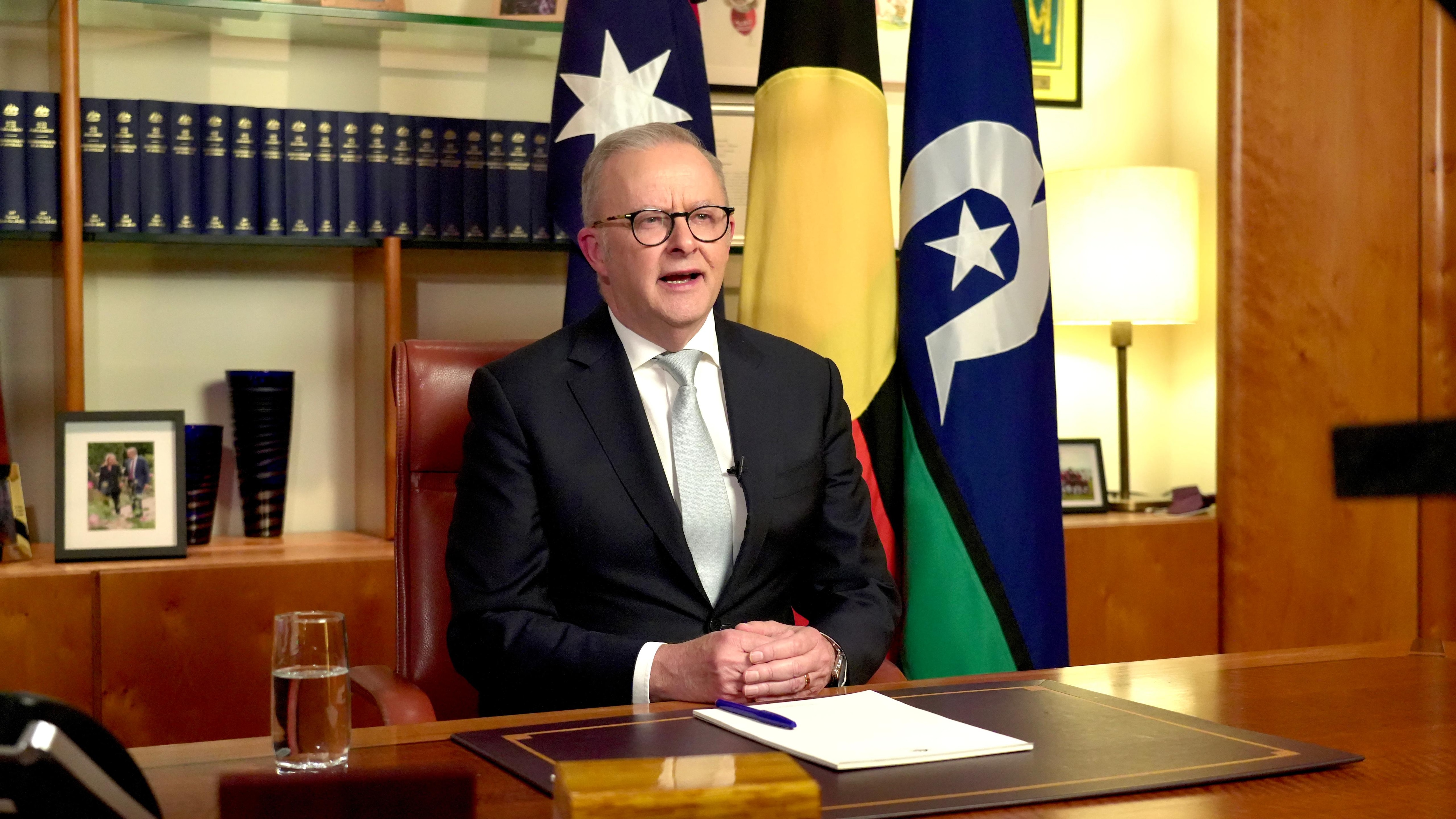 A man in a suit sits at a desk and reads a teleprompter, looking in to a camera.