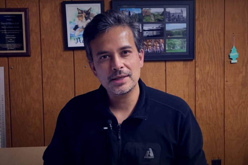 A man wearing a blue shirt sits in front of a wooden wall.