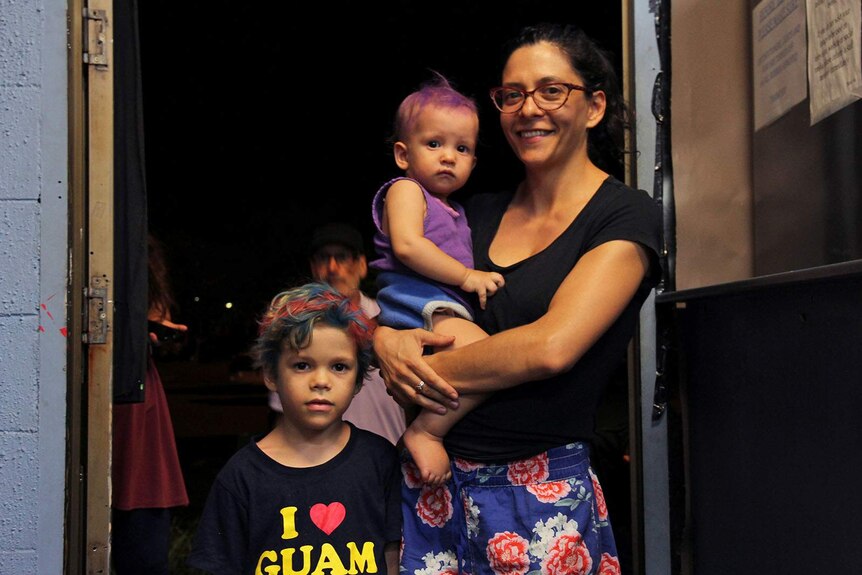 A woman stands with her two young children in a doorway.