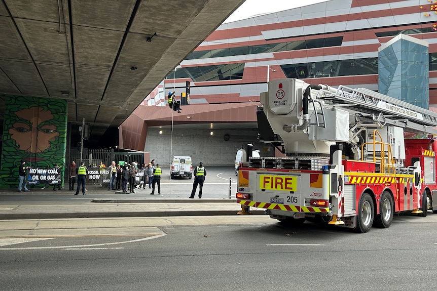 A person abseiling from a bridge with people holding signs in the background and a fire truck on the right