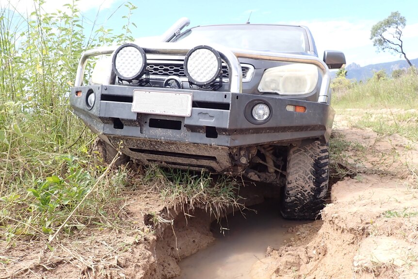 A 4WD leans to one side where a tyre is submerged in a deep pot hole on a dirt road.