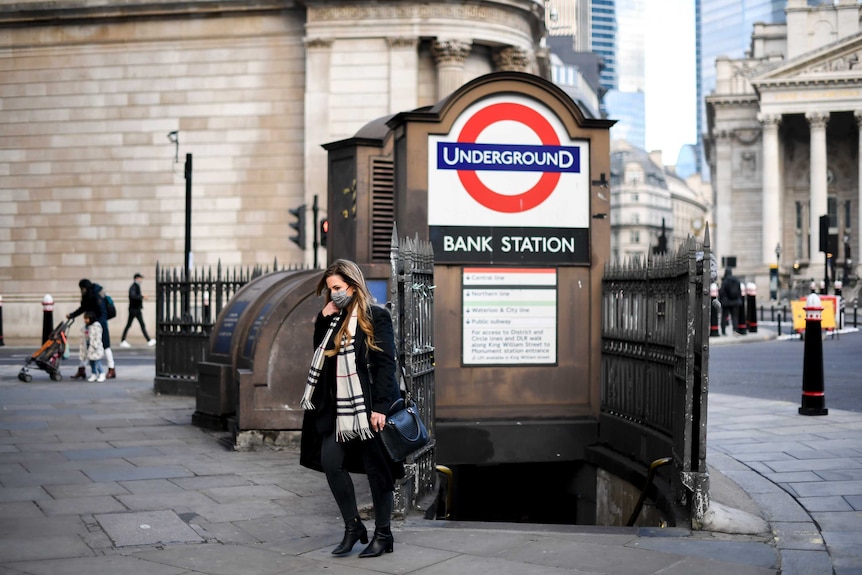 A woman wearing a face mask, scarf and coat emerges from a set of stairs. A sign says UNDERGROUD: Bank Station