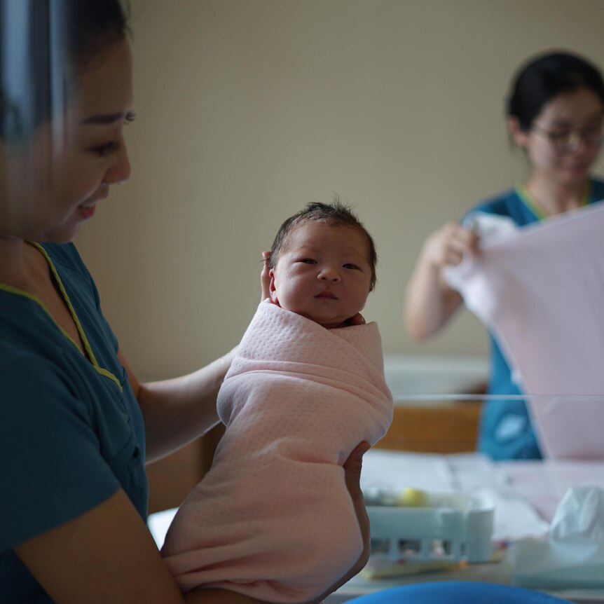 A midwife holds a baby up to the camera, another helps in the background folding a sheet.