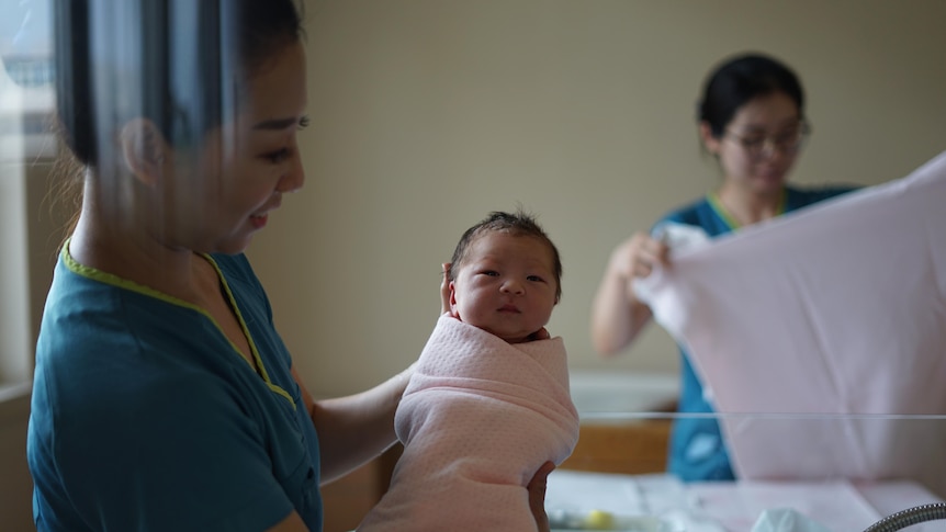 A midwife holds a baby up to the camera, another helps in the background folding a sheet.