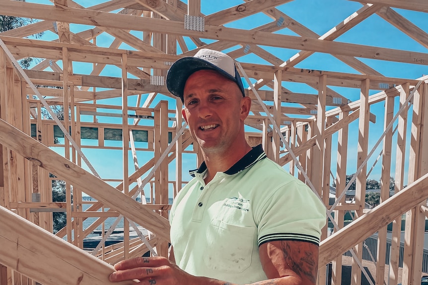 A man with tattoos on his arms wears a high visibility top and stands in a construction site smiling at the camera
