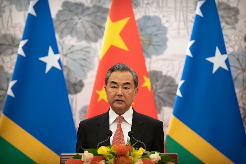 Wang Yi stands at a lectern in front of a Chinese flag and two flags of the Solomon Islands. 