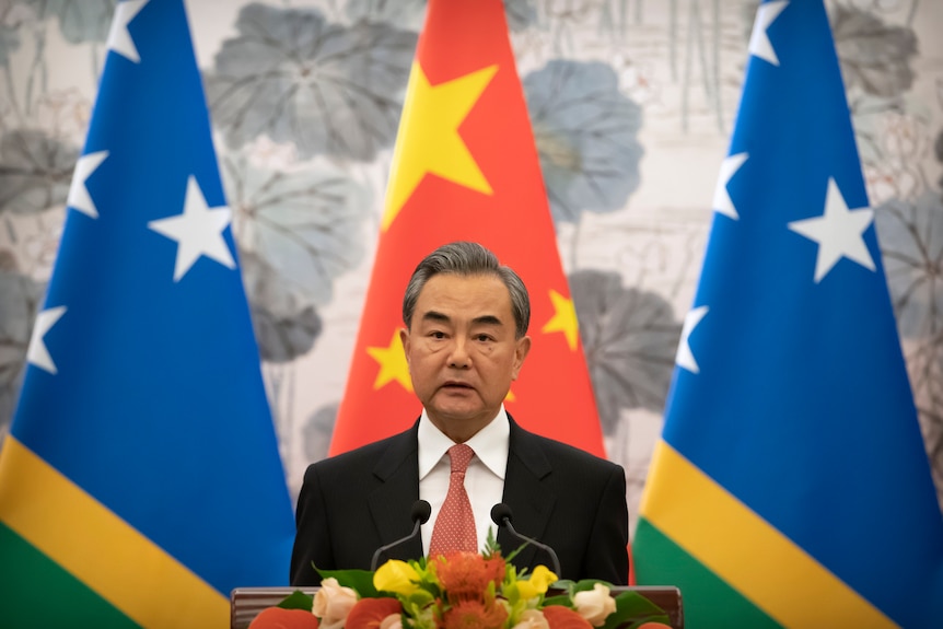 Wang Yi stând în fața unui amvon, în fața unui steag chinez și a două steag ale Insulelor Solomon. 