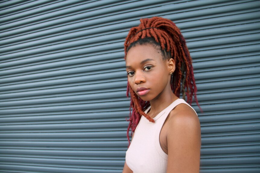 Ani, who has dyed red dreadlocks, looks at the camera in a white tank top.