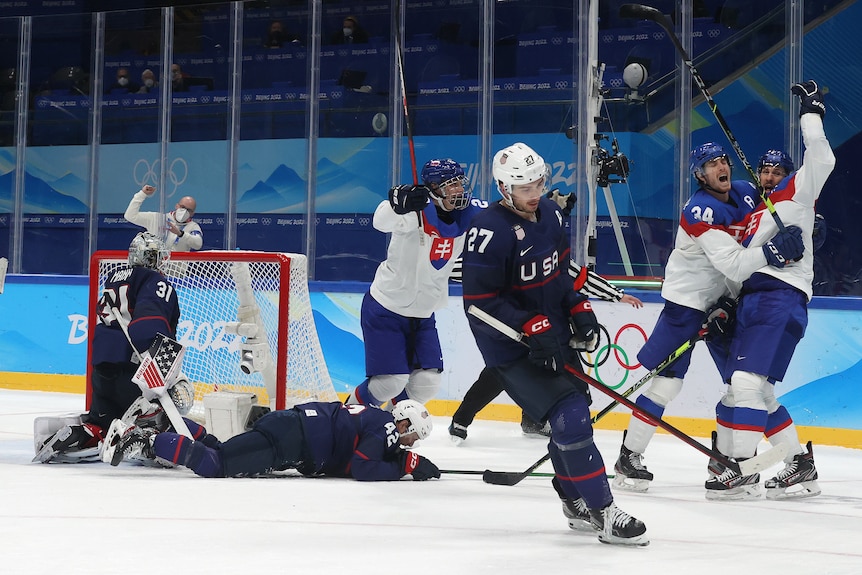 Slovenskí hokejisti oslavujú pri pohári, zatiaľ čo zlomení hráči Spojených štátov ležia po góle na ľade.