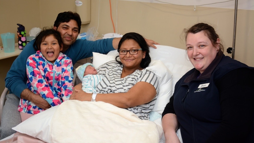 A couple with their newborn baby and daughter in a hospital room, with the nurse that helped with the birth on roadside.