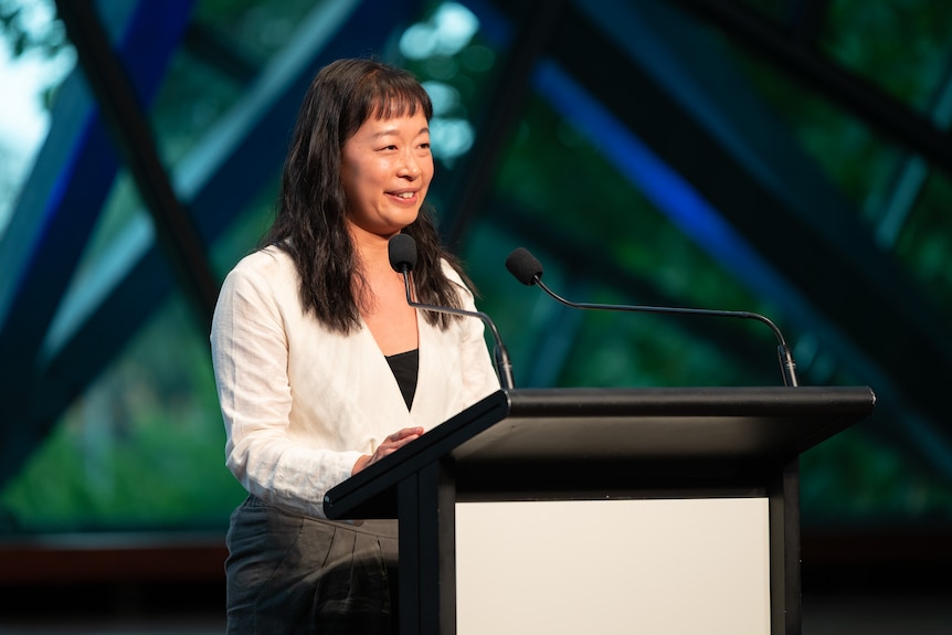 36-year-old Chinese Australian woman with long black hair smiling, standing at podium.