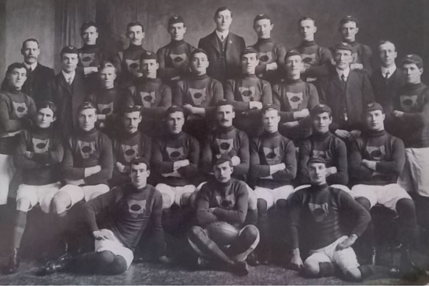 A black and white photo shows men with folded arms from an AFL team posing for a photograph.