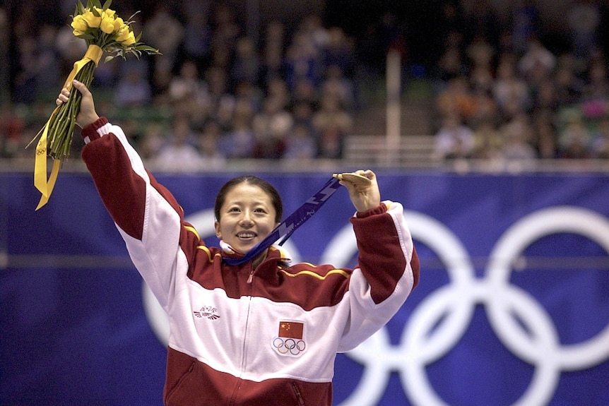 Speed skater stnad on top of the podium with her gold medal and flowers.