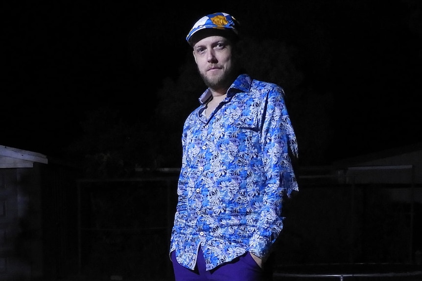 Un hombre parado afuera por la noche con un sombrero plano, camisa azul y pantalones morados
