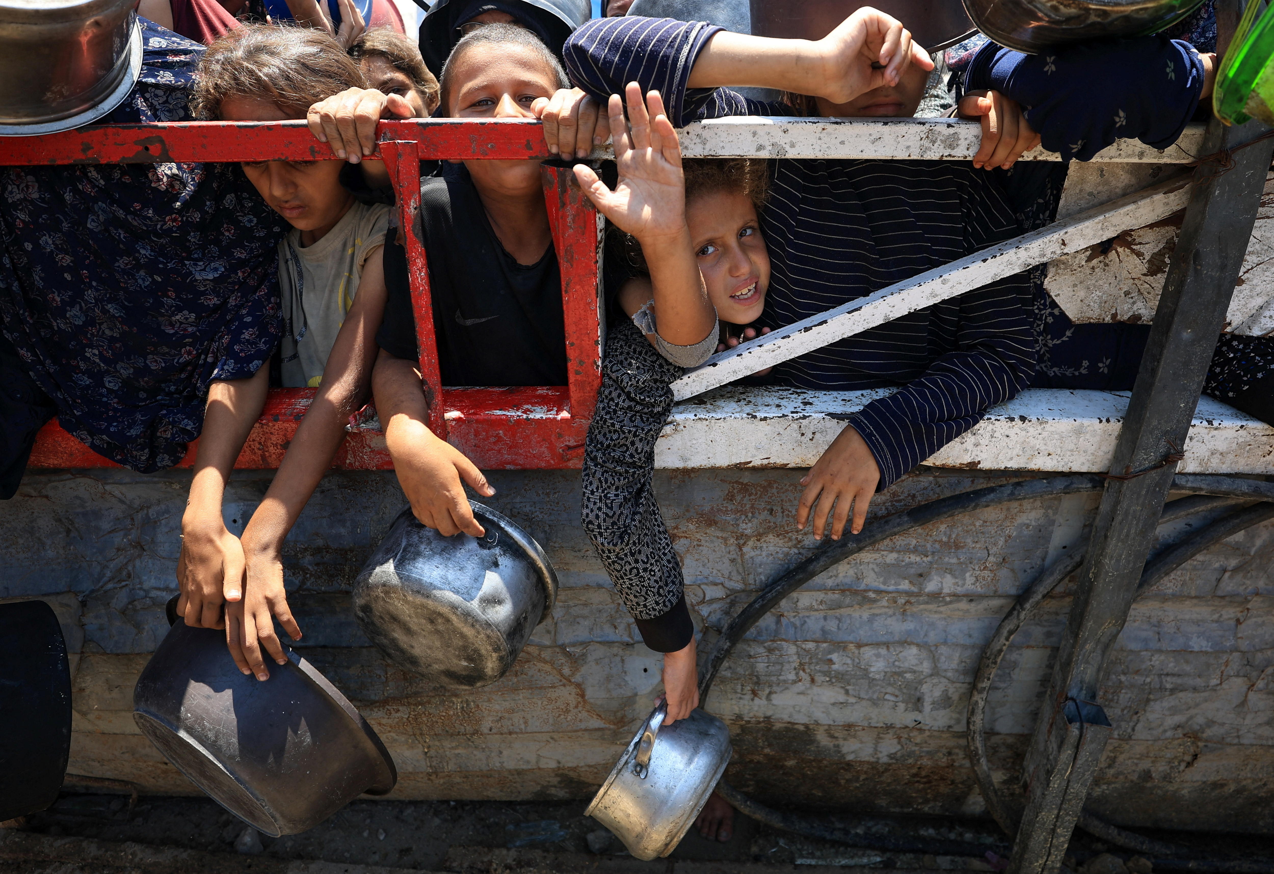 Children pressed against a metal railing holding out food containers in their hands