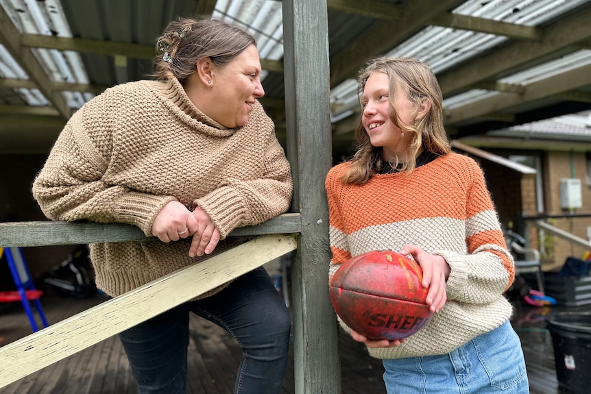 A woman leaning on a verandah and a young woman leaning against a nearby post, holding an AFL football