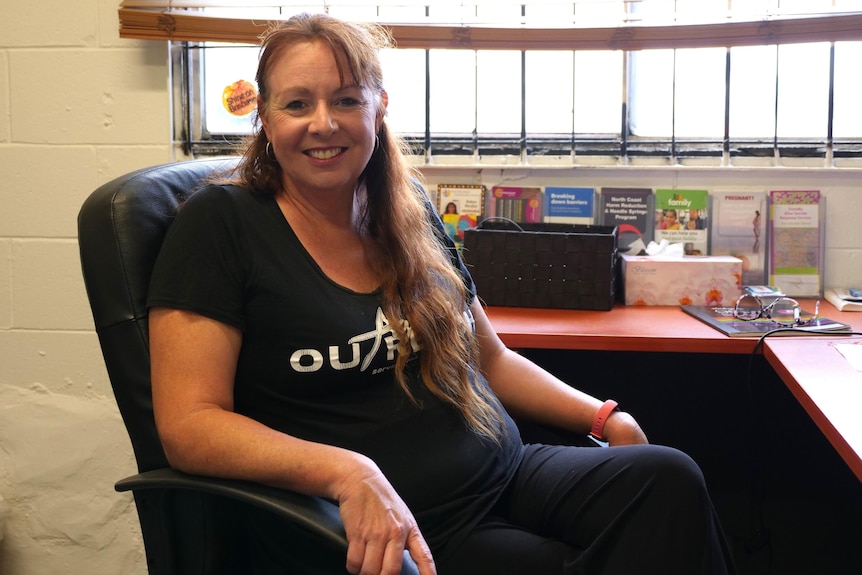 A woman in an Agape Outreach t-shirt smiles at the camera while seated at a desk in an austere office.