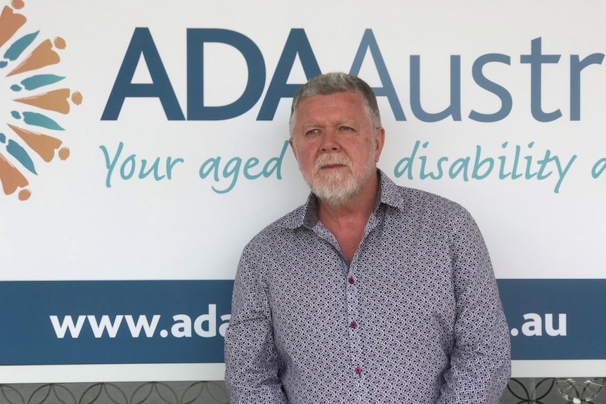 A man with a beard and grey hair standing front of an ADA Australia sign. 