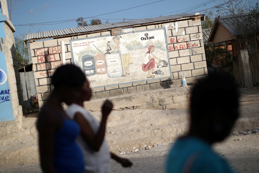 Women in Haiti walk past an Oxfam sign on a dusty road