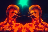 A symmetrical image of David Bowie with a blonde mullet and patterned turtle neck singing into a mic under red and green lights.