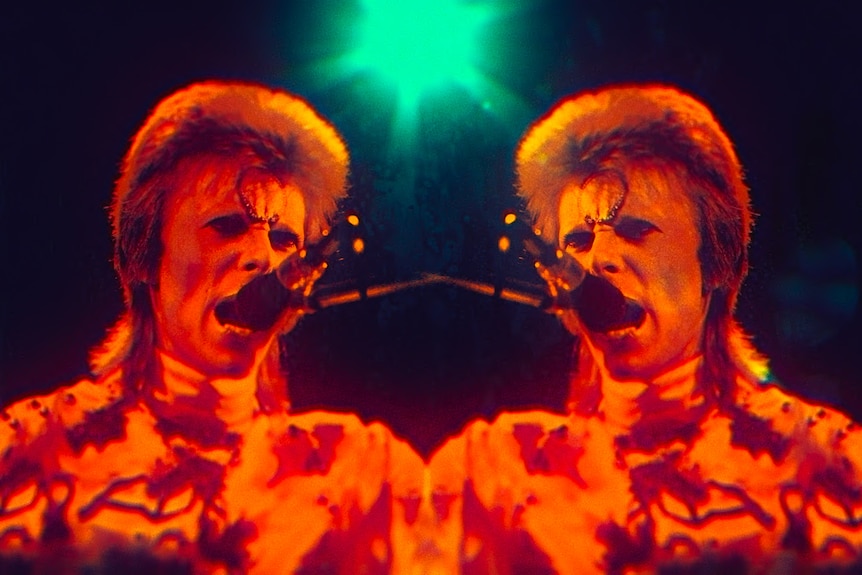 A symmetrical image of David Bowie with a blonde mullet and patterned turtle neck singing into a mic under red and green lights.