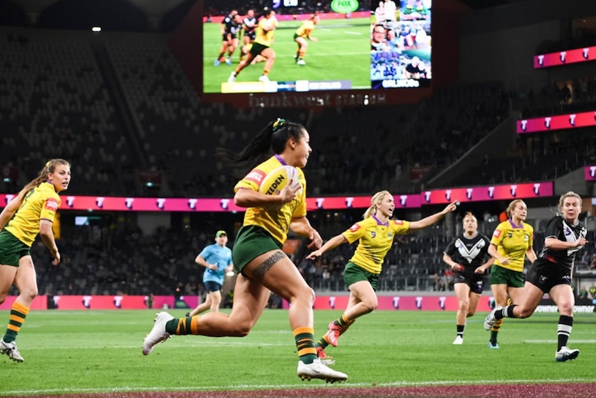 Woman runs the field holding football in yellow and green Jillaroos uniform surrounded by plaers