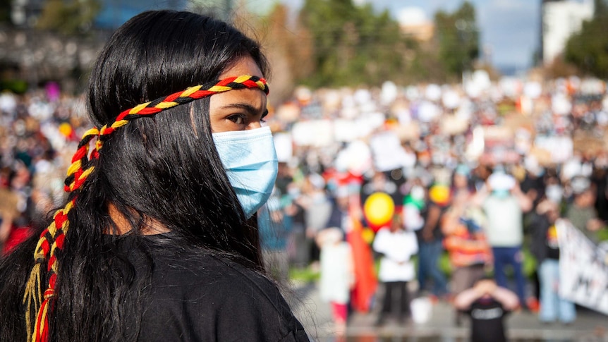 A woman with a red, black and yellow Aboriginal flag headband and a facemask, with the Black Lives Matter protest behind.