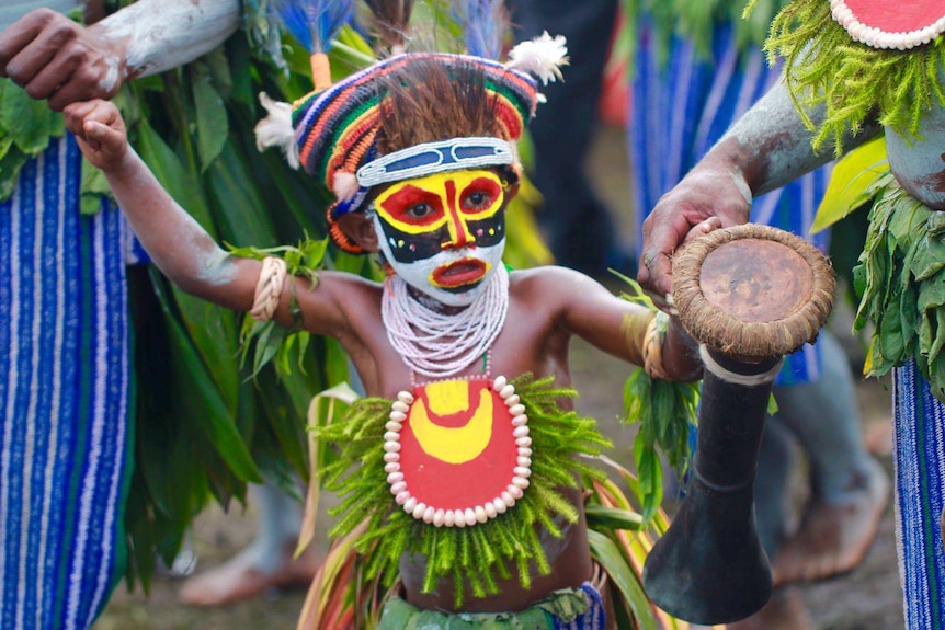A child in red, white, yellow and black facepaint holding a Kundu drum at the 2016 Goroka Show, Papua New Guinea.