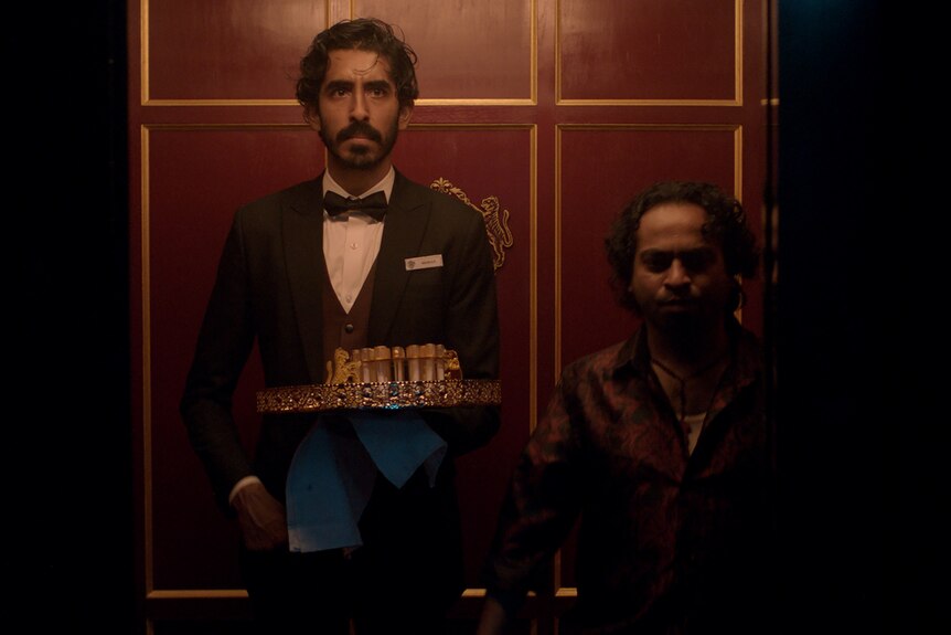 A film still of Dev Patel and Pitobash, standing together in an old-fashioned lift. Patel has a tray of drinks in one hand.