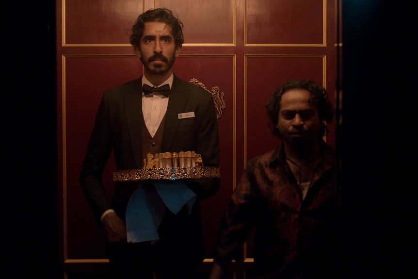 A film still of Dev Patel and Pitobash, standing together in an old-fashioned lift. Patel has a tray of drinks in one hand.