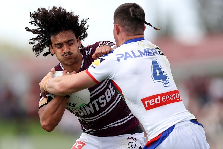 a Manly NRL player holds the ball as he is tackled across his chest by a Newcastle opponent.