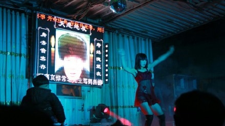 A stripper performs at a funeral in China. There is an image of a man's face with a condolence message on the screen behind her.