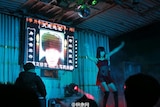 A stripper performs at a funeral in China. There is an image of a man's face with a condolence message on the screen behind her.