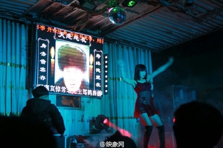 A stripper performs at a funeral in China. There is an image of a man's face with a condolence message on the screen behind her.