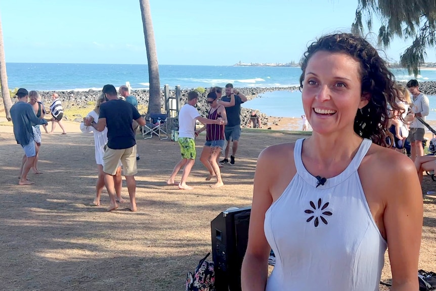 A smiling, curly-haired woman looks just past the camera as a group dances near a beach behind her.