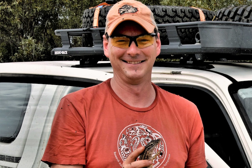 Man wearing spectacles standing in front of a four-wheel drive, holding a reptile