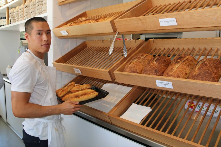 Baker Ryan Chu stands with a tray of bread rolls next to shelves containing more bread rolls.