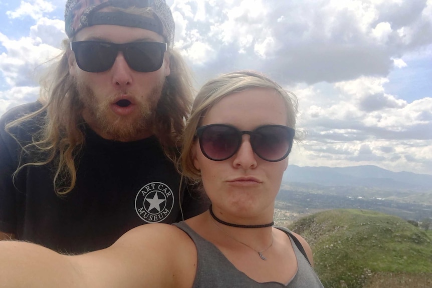 A man with long hair and blonde woman, both wearing sunglasses, taking a selife on a mountain.