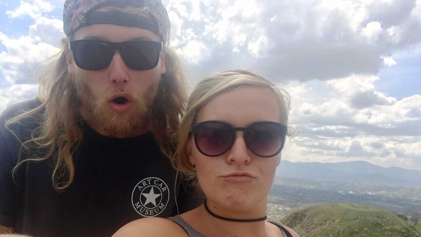A man with long hair and blonde woman, both wearing sunglasses, taking a selife on a mountain.