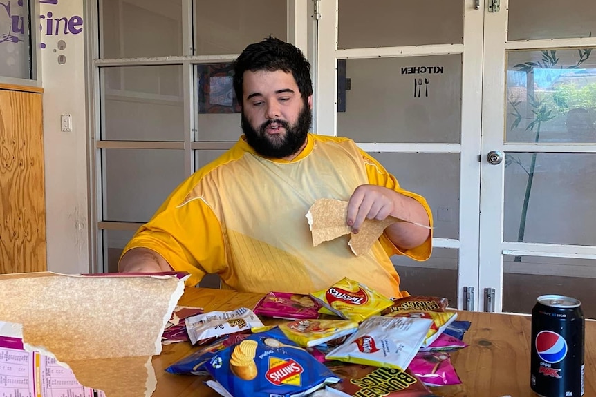 A young heavy set man with a beard. He's tearing paper and chip packets