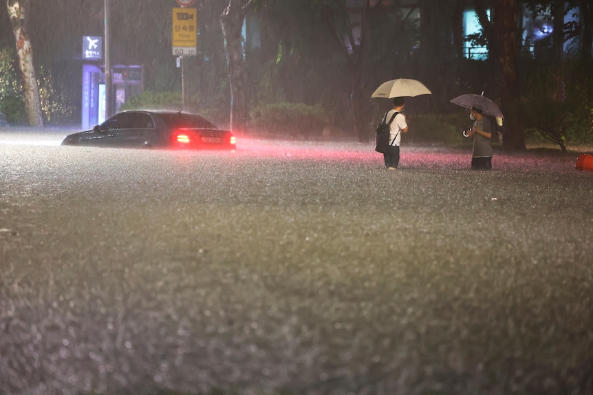 Two men wade through thigh-high water on a flooded road with a car nearby as it rains.